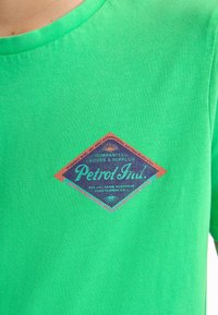 Groene katoenen t-shirt met een diamantvormige print in oranje en blauw, met de tekst "Petrol Intl." Gestructureerde stof met een ronde hals.