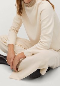 Beżowy sweter o wysokiej stójce, zestawiony z jasnymi, szerokimi spodniami. Modelka siedzi, prezentując luźny krój i fakturę.