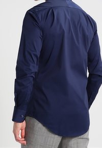 Navyblaue Langarmshirt mit glatter Textur, geknöpften Manschetten und abgerundetem Saum. Sichtbare Nähte und subtile Schulterdetails.