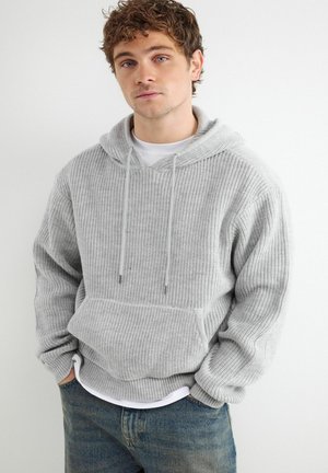 Mladý muž s kučeravými vlasmi, oblečený v svetlosivej pletenej mikine s kapucňou a modrých džínsach, stojí so zloženými rukami v kapse pred neutrálnym pozadím.