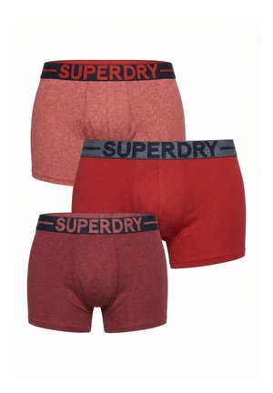 Tre boxer rosso da uomo in diverse tonalità, ciascuno con una fascia nera in vita con il nome del marchio "SUPERDRY" in lettere grasse.