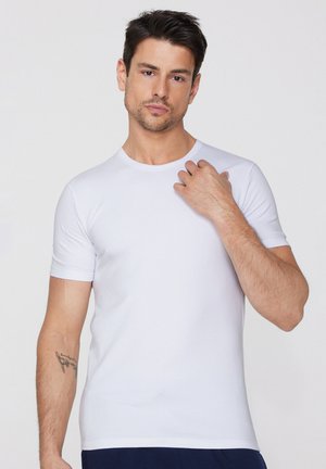 Mann mit kurzem dunklem Haar, der ein einfaches weißes kurzärmeliges T-Shirt trägt und mit seiner rechten Hand seine Brust berührt, neutraler Hintergrund.