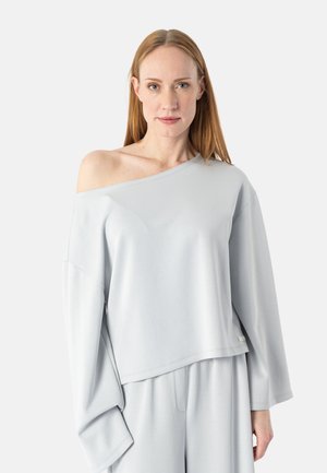 Kvinne med rett rødt hår iført løs, lys grå skulderfri topp og matchende vide bukser mot en ensfarget bakgrunn.