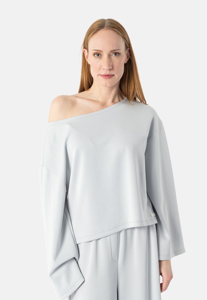 Donna con capelli rossi lisci che indossa un top largo grigio chiaro scivolato sulle spalle e pantaloni abbinati a gamba larga su sfondo neutro.