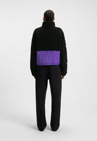 Giacca in fleece nera con un pannello in nylon viola in vita. Collo alto, vestibilità comoda e maniche lunghe. Abbinata a pantaloni neri.
