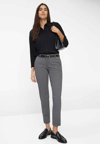 Schwarz langärmliges Hemd, in graue karierte Hose gesteckt, mit einem schwarzen Gürtel gesichert. Schwarze Slipper und eine schwarze Handtasche runden das Outfit ab.