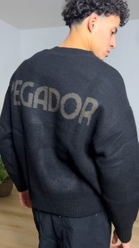 Black knit sweater with a loose fit, featuring "VEGADOR" in a contrasting gray on the back. Ribbed hem and cuffs, soft texture.