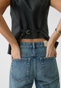 Czarny top bez rękawów z fakturowanym wzorem w kropki i detalem w postaci bocznego paska, zestawiony z niebieskimi dżinsami z tylnymi kieszeniami.