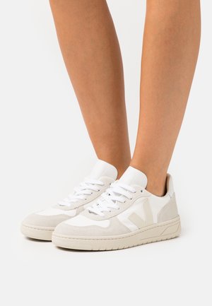 Hvide og beige sneakers med lavt skaft og snørebånd, båret på bare ben mod en ensfarvet hvid baggrund.