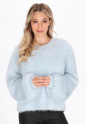 Junge Frau mit langen blonden Haaren, die einen hellblauen, flauschigen Pullover mit Perlenverzierungen trägt und mit gefalteten Händen posiert.