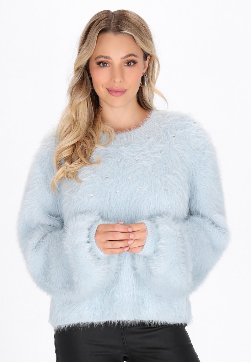 Junge Frau mit langen blonden Haaren, die einen hellblauen, flauschigen Pullover mit Perlenverzierungen trägt und mit gefalteten Händen posiert.