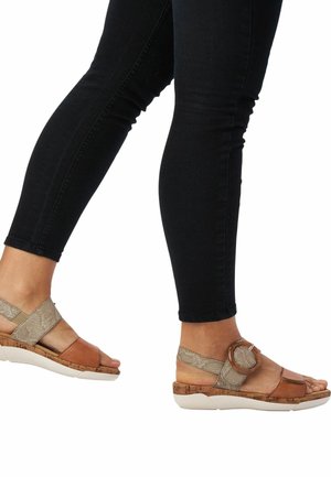 Sandales en cuir marron avec une semelle en liège, présentant une sangle accentuée en or texturé et une boucle métallique ronde, portées avec un jean noir.