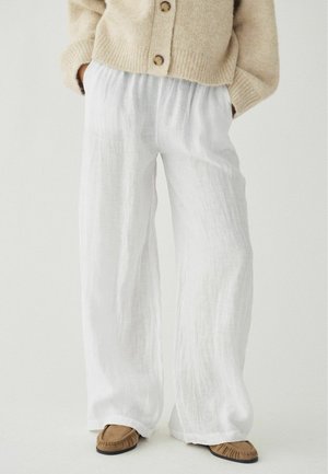 Inimene, kes kannab lahtiseid valgeid linaseid pükse, beežist nuustikutega kootud kampsunit, millel on nööbid, ja pruuni mokassiinide tüüpi jalanõusid, käed taskutes.