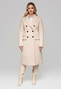 Mujer vestida con un abrigo beige de doble botonadura con botones negros, camisa blanca y botas altas a la rodilla a juego, de pie contra un fondo gris.