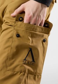 Braune Cargo-Hosen-Details mit einer Tasche mit schwarzen Druckknöpfen und einem großen schwarzen "SLADE"-Logo. Der Stoff scheint robust und mit einer glatten Textur versehen zu sein.