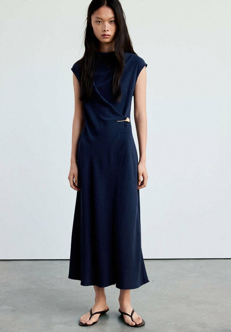 Femme debout portant une robe portefeuille sans manches bleu marine maintenue par une épingle dorée et des tongs noires sur un sol gris.