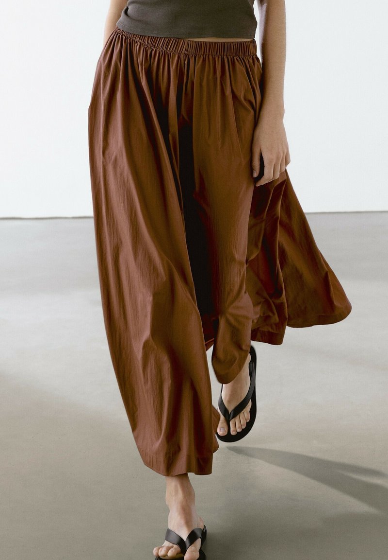 Personne marchant portant un pantalon marron ample à jambes larges avec une taille élastique et des tongs noires sur un sol lisse.