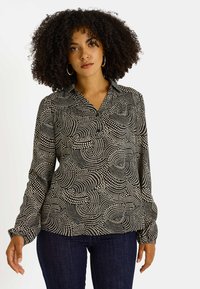 Blusa con estampado en blanco y negro de círculos ondulados, con cuello, botonadura y mangas largas, combinada con vaqueros azul oscuro.