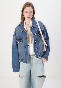 Veste en denim bleu moyen avec des détails usés, grand col et finitions en métal argenté. Portée par-dessus une chemise blanche avec des accents en paillettes et un jean léger bleu déchiré.