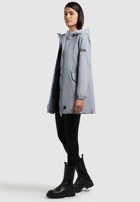 Frau trägt eine hellgraue Kapuzen-Regenjacke, schwarze Leggings und klobige schwarze Stiefel, steht seitlich auf einem schlichten hellen Hintergrund.