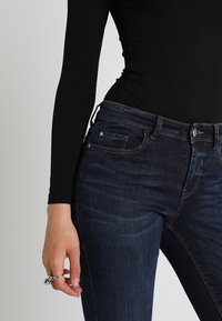 Kvinna med mörkblå denimjeans och en figursydd svart långärmad tröja, med en silverring på sin vänstra hand.