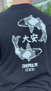 Musta t-paita, jossa on valkoinen graafinen kuva kahdesta koi-kalasta ja teksti "OSAKA" japanilaisilla merkeillä alapuolella. Pehmeä puuvillamateriaali, rento muotoilu.