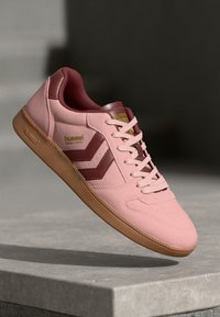Sneaker rosa in suede con accenti marroni, design con lacci, suola in gomma e dettagli con logo di lato. Presenta una texture perforata sulla parte superiore.