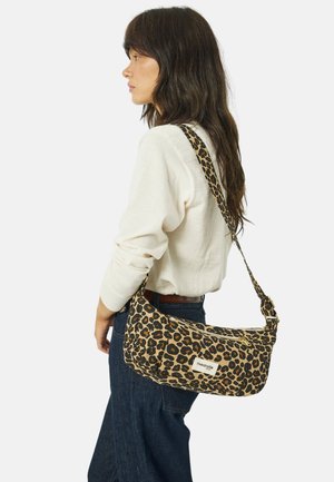 Woman in white sweater and dark jeans carrying leopard print shoulder bag with rivedroite label, facing left in profile.