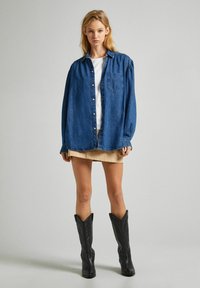 Camicia in denim oversize con chiusura a bottoni, taschino sul petto, e spalle arricciate, indossata sopra una maglietta bianca e una gonna beige, abbinata a stivali neri.