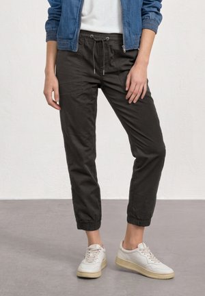 Person trägt schwarze Jogginghosen mit Kordelzug, weiße Sneakers, ein weißes Hemd und eine teilweise gezippte blaue Jeansjacke und steht auf grauem Boden.