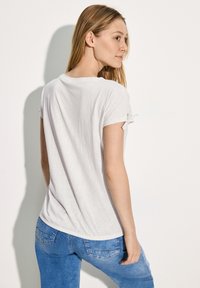 Weißes T-Shirt mit kurzen Ärmeln und strukturiertem Muster, mit aufgerollten Ärmeln und abgerundetem Saum, kombiniert mit hellblauen Jeans.