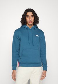 Tīklas kapuces džemperis mīkstā audumā ar aukliņām, kenguru kabatu un logo detalizāciju uz krūtīm. Apvienots ar teksturētām gaišām biksēm.