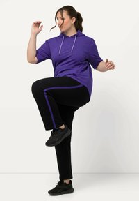 Paarse hoodie met korte mouwen en een trekkoord, gecombineerd met zwarte broek met een paarse zijstreep, en zwarte sportschoenen.