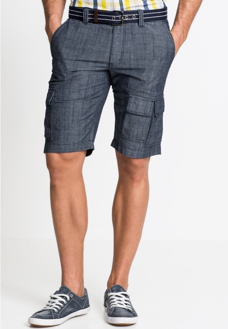 I pantaloni cargo in denim blu scuro presentano molteplici tasche, una parte anteriore piatta e un orlo risvolto. Abbinati a sneakers in tela grigie.