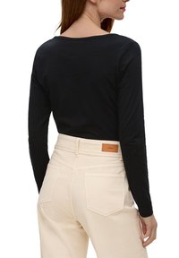 Schwarzes Langarmoberteil kombiniert mit hellbeigen High-Waist-Jeans. Auffälliges braunes Lederpatch auf der Rückseite der Jeans. Glatte Textur.