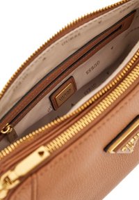 Interno di una borsa in pelle sintetica marrone con cerniera oro, dotato di fodera in tessuto contrassegnata dal marchio "Guess" e un piccolo tag con il logo.