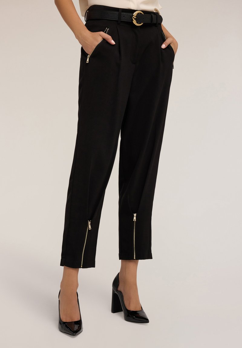 Zwarte tapered broek met ritszakken en ritssluitingen bij de pijpen. Voorzien van een zwarte riem met een gouden ronde gesp. Gladde stoffen structuur.