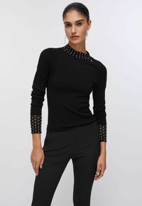 Pull noir en tissu maille, avec un col rond clouté et des poignets ornés. Coupe slim, manches longues.