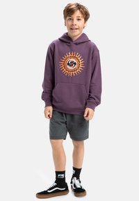 Lila hoodie med en solgrafik i orange, känguruficka, grå shorts, svart-vita sneakers och svarta strumpor.