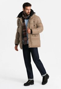 Chaqueta acolchada beige con cuello negro, detalles de botones, llevada sobre un suéter azul claro. Jeans oscuros y botas negras con cordones y acentos dorados.