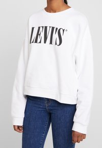 Biała, skrócona bluza dresowa z miękkiego materiału, z dużym czarnym logo "LEVI'S" na klatce piersiowej, z prążkowanymi mankietami i dołem.