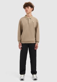 Beige poloshirt met lange mouwen, een kraag en geborduurde details, gecombineerd met zwarte broek en zilveren sneakers. Glad textuur van de stof.
