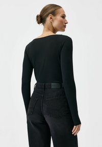 Kobieta z kokiem na głowie, ubrana w czarną bluzkę z długim rękawem wpuszczoną do czarnych, wysokich dżinsów, stojąca na tle jednolitego tła.