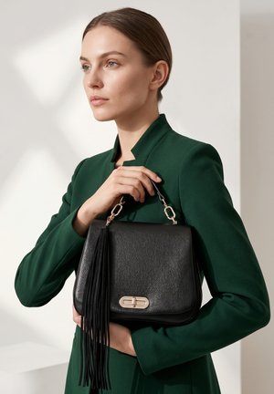 Frau in grünem Blazer hält schwarze Lederhandtasche mit goldener Schnalle und langem Quaste vor neutralem Hintergrund.