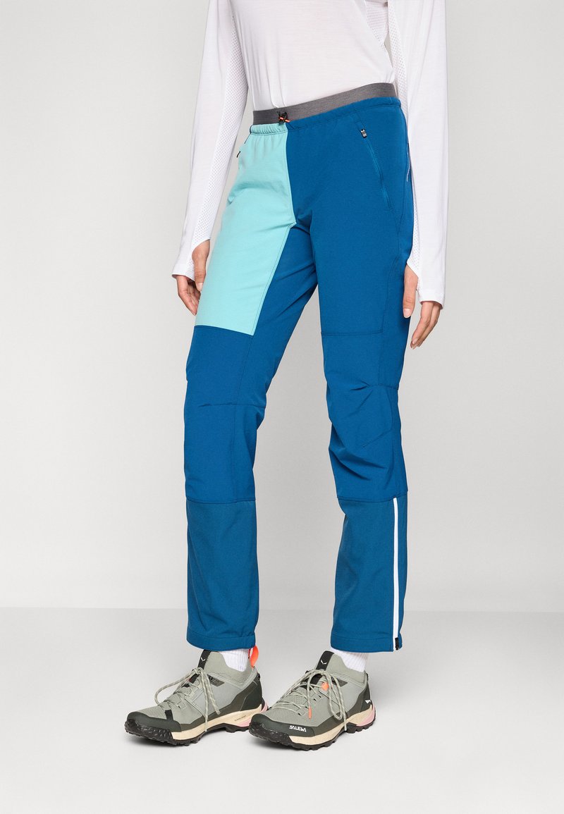Blaue und türkisfarbene Outdoor-Hosen mit tailliertem Design, ausgestattet mit Einsätzen, elastischem Bund und Reißverschlusstaschen. Tragen mit Wanderschuhen.