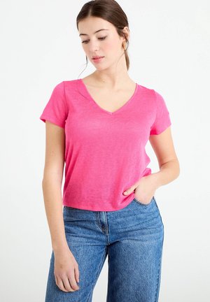 Donna con capelli castani, indossa una maglietta rosa a V e jeans blu, una mano nella tasca, guarda verso il basso su sfondo bianco.