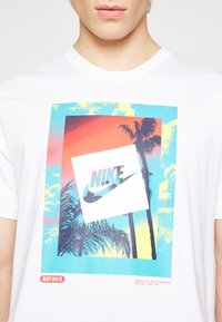 Lähikuva valkoisesta t-paidasta, jossa on värikäs auringonlasku, palmupuita ja vinosti oleva Nike-logo keskellä, tekstinä "Just Do It." alla.