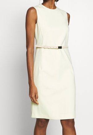 Bílé šaty bez rukávů po kolena se štíhlým světlým páskem, které nosí stojící osoba na jednobarevném pozadí.