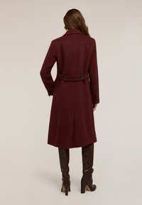 Cabotto in lana bordeaux con vita aderente, caratterizzato da un design doppiopetto e dettagli con bottoni, che arriva a metà polpaccio.