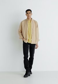 Beige overhemdd met knopen over een gele T-shirt, gecombineerd met zwarte jeans en zwarte laarzen. Simpel, relaxte pasvorm; eenvoudige ontwerp zonder patronen.